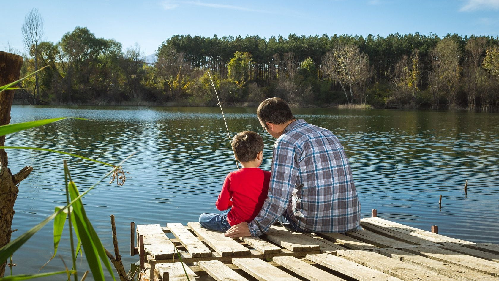 Man and child fishing from a wooden dock on a calm lake with trees in the background